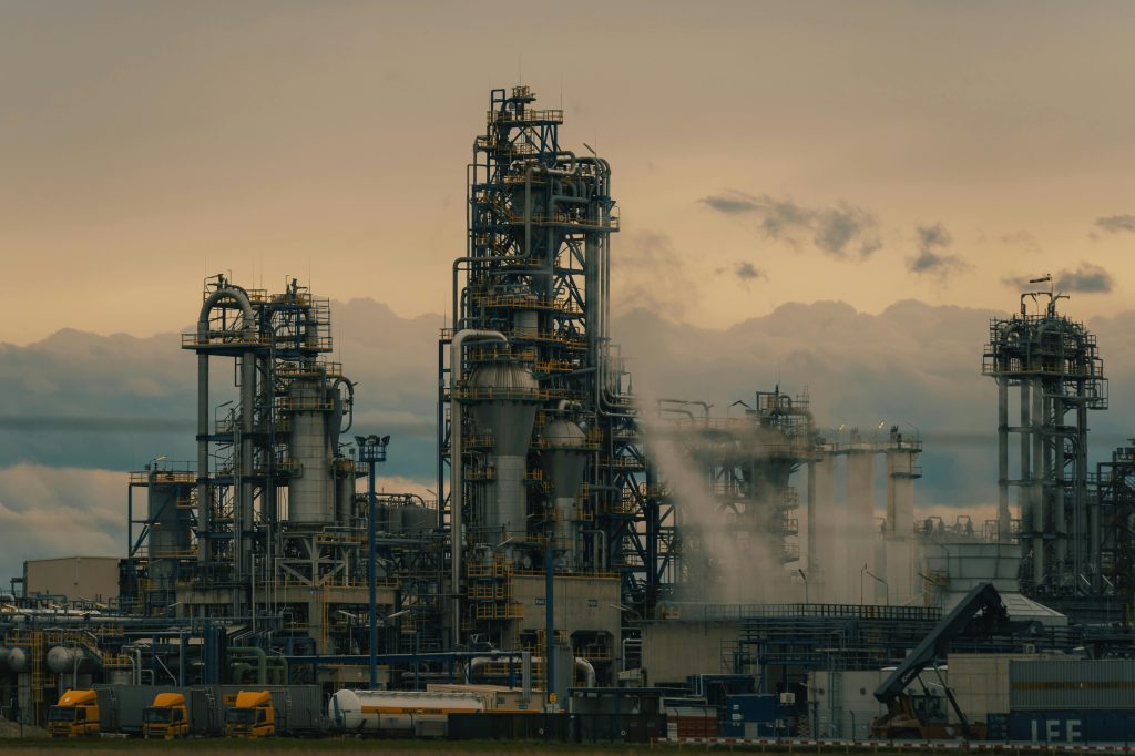 An industrial landscape image featuring a large oil refinery with storage tanks, pipes, and smokestacks.